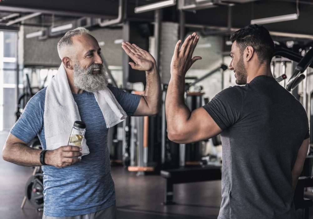 Give me five. Joyful men are standing in sport club and enjoying friendly conversation. Senior pensioner is holding bottle of water in his hand